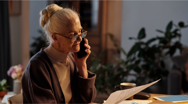 An older adult sits at a desk, holding a phone and reviewing paperwork as they calculate health care costs in retirement.